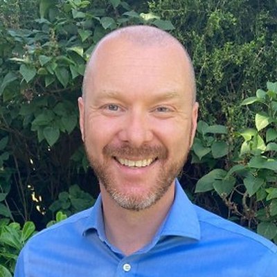 Chris Wyle, smiling, in a blue shirt in front of green foliage